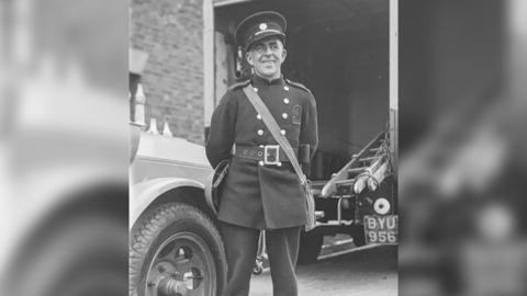 A man posing for the camera in a button up jacket and hat of the firefighter's uniform. He is smiling at the camera and behind him there is an old vehicle with a ladder on the back of it.