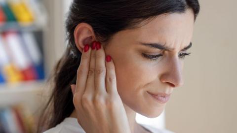 A close-up of a person’s face. They are frowning and have their fingers pressed against the middle of their ear.