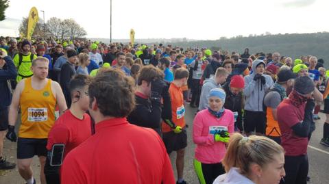 A large group of runners in a variety of brightly colored vests, sports tops and leggings huddle together at the start line.