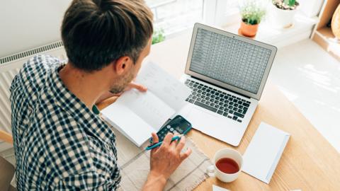 Man using calculator and laptop computer to calculate numbers, focused on work and appears to be working on financial or accounting task.