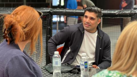 Muhammad is sat down at a metallic table looking at a woman sitting opposite him. He is wearing a blue jacket over a white T-shirt. He's leaning a bit to his right. Behind him is a padded fence for a children's play area as a couple of people queue to enter the venue.