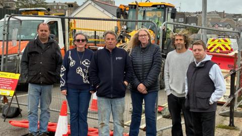 Six traders stand in front of the road closure in Penzance with works site vehicles behind them. 