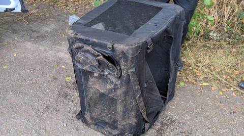 A black container with netted sides which is on the ground. It is wet and covered in sand and dirt.