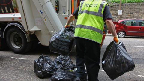 A person wearing a yellow high-vis vest carries bin bags in either hands towards the rear of a bin lorry parked on a road. His body and legs are in shot but his head is not. There are a small pile of bin bags at his feet.