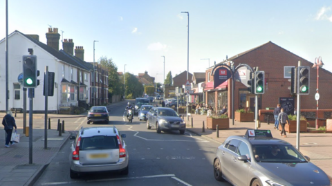 Rainham town centre. The road has a number of cars on it and three traffic lights all showing green lights. A number of buildings are in the background on either side of the road.