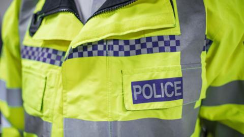 A close‑up of a bright yellow high‑visibility police jacket with reflective silver stripes and a blue‑and‑white check pattern, featuring a bold blue patch reading “POLICE” on the front pocket.