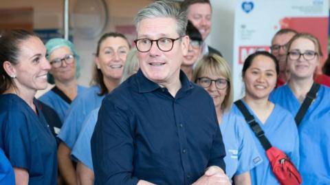 Sir Keir Starmer, wearing a navy blue shirt, stands with his hands clapsed, with NHS staff in blue scrubs stand behind him