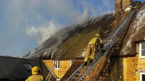 House with thatched roof - several ladders are against the roof with two firefighters up a ladder and another at the bottom. Smoke is pouring from the roof of the home.