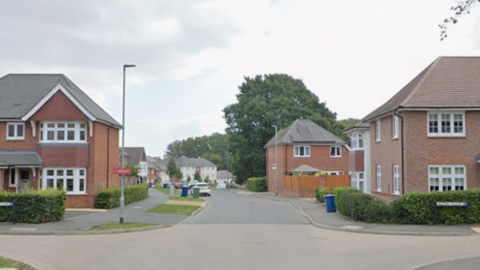 A street view picture of a modern residential street with houses along both sides, cars parked on the left and several homes with wheelie bins outside.