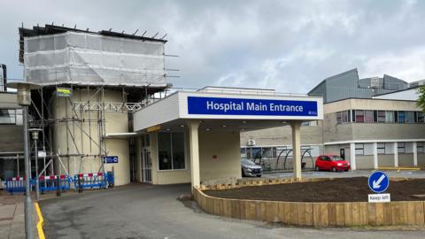 Guernsey hospital main entrance with blue signage, a covered drop‑off area and scaffolding wrapped in white sheeting on an adjacent building under grey, overcast skies.