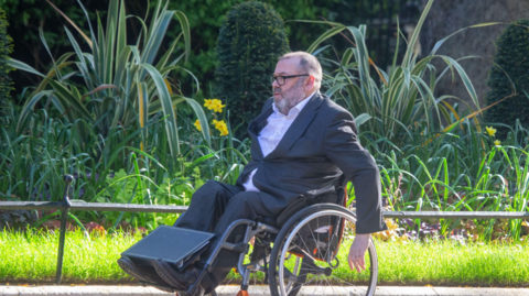 David Williams, Permanent Under-Secretary of State for Defence of the United Kingdom, is seen arriving at 10 Downing Street, in April 2025. He is a wheelchair user and wears a dark grey suit with a white, open-necked shirt, and glasses. 