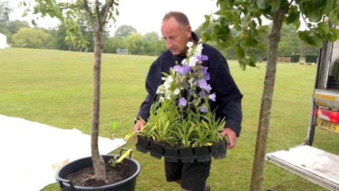 A man in a dark top walks between two trees carrying pots of flowers.
