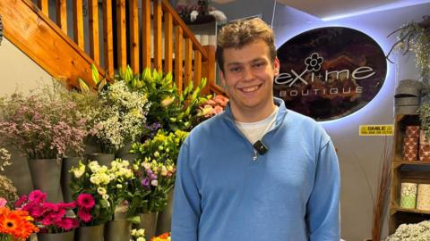 Jamie is wearing a blue quarter zip sweater. He is smiling and behind him is an array of flowers and plants.