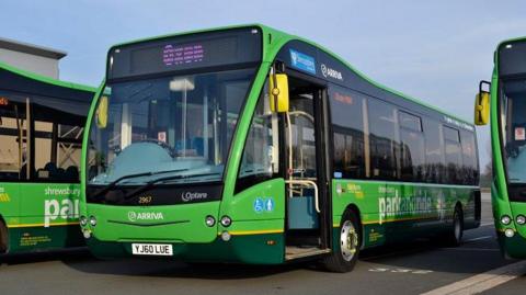 A green Arriva bus with the words Shrewsbury Park and Ride on the side, parked alongside two other buses and bathed in sunlight.