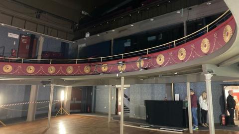 interior of two-tiered audience area with balcony adorned by red and gold decor and cordoned off area on ground floor