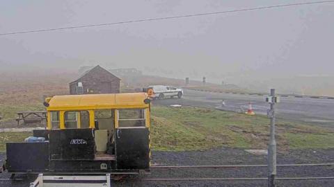 The Mountain Road at the Bungalow. There is a small train engine with a yellow roof on the tram tracks in the foreground, and a white van across the road.