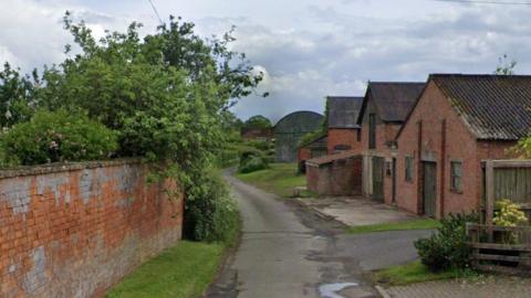 A view of Monks Lane. There are no cars parked along the rural lane which is flanked by farm buildings and walls made of red brick.