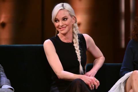 Anna Faris smiles at someone off camera, she has long blonde hair in a side plait. She wears a sleeveless black top.
