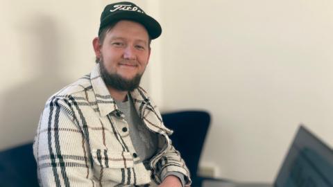 Mr Digby, digital inclusion manager, is facing the camera and smiling. He is a white man with brown hair and a brown beard. He is wearing a black cap, a grey T-shirt, and a cream jacket with black and brown tartan stripes.