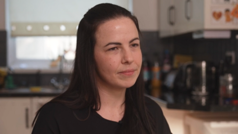 Close up of a woman with long dark hair, wearing a black t shirt. She is looking off to the side with a serious expression.