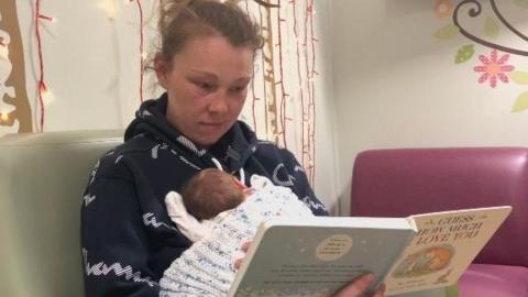 A woman in a blue top is holding a baby in front of her and holding a book in her other hand. She is sitting on a green chair with a purple sofa in the background and patterns and lights on the walls.