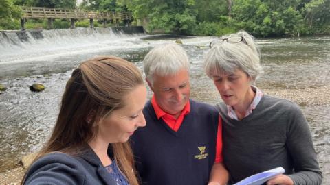 Left to right Derby North MP Catherine Atkinson, John Rickard and Joanna West are looking at plans for turbine. They are stood on a bank looking at the weir.