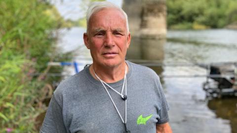 A man with white hair, wearing a grey T-shirt, stands in front of a river 