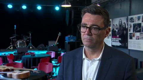 Andy Burnham. He has dark hair, wearing glasses, a navy blazer and white shirt.