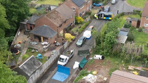 A drone shot above several homes and gardens - a residential road running through the middle which has a forensics tent, police van and sheets up to cover areas.