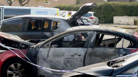 Several burnt cars in a row with police tape over them. The cars are all charred. There is a white "scientific services" van in the background.