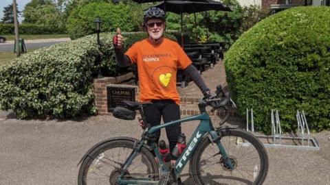 A man in an orange t-shirt and long black sleeve shirt is standing by a bicycle. He is wearing a helmet and a pair of sunglasses.