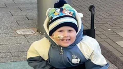 A boy with a blue and white coat, blue gloves an a hat with Bluey on it sat in a wheelchair on a pavement with a blue blanket on his lower half.