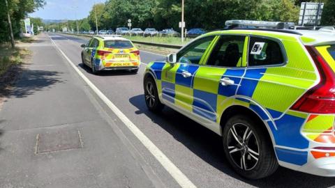 Two police cars are parked at the side of a road with no other traffic on it. On the opposite carriageway cars can be seen in a long line.