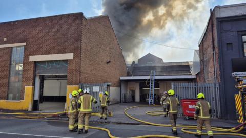 Seven firefighters dressed in brown firefighting uniforms with yellow helmets work at the scene of a fire. Smoke is billowing into the sky behind a building in the background. Yellow hoses from a fire truck on the right can be seen snaking along the ground and a ladder has been placed against a roof in the background.