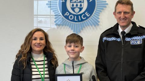 A boy holding framed award looks at the camera with a woman and police man