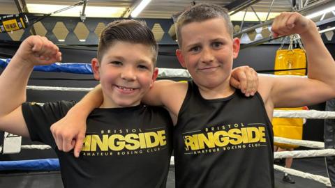 Photo of Jake and Tommy in front of the boxing ring with their arms round each other. They are smiling and wearing black Bristol Ringside tops.