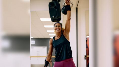 A person wearing a black vest top and purple leggings holds a kettle bell in the air and smiles. In the background you can see a gym studio with a speaker hanging from the ceiling.