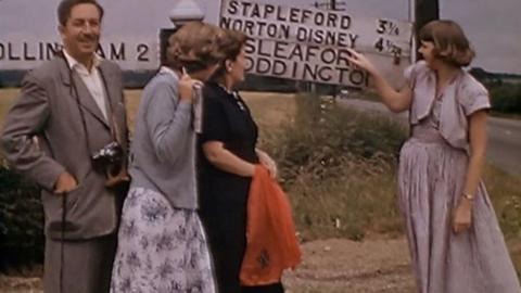 Walt Disney with a camera and female members of his family standing by a road sign to Norton Disney with one of the women pointing at the sign