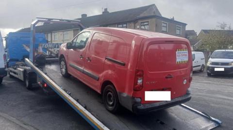 A red van with a "fly tipping" label on the back is loaded onto a pick-up truck.