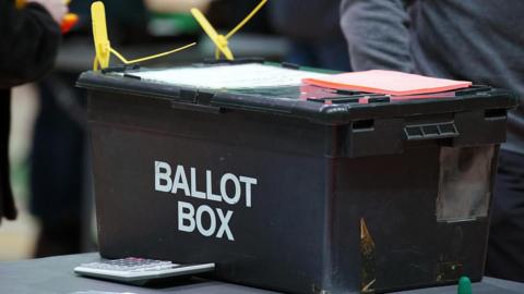 Black ballot box with white writing on it. It is locked with cable ties and there are papers on top of it and a calculator in front.