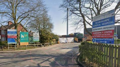 The entrance to a hospital with fencing either side of a road going into it. Either side of the fencing you can see signs saying 'NHS Scunthorpe General Hospital, Outpatients Entrance, Emergency Centre'.