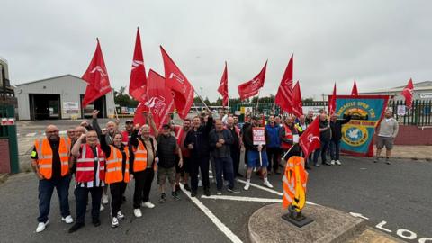 Crowds of men and women, some wearing orange hi-vis jackets, stand outside a bus depot, holding red Unite flags. 