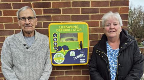 Man with grey hair and glasses and women with black coat and blue top stood side by side next to a defibrillator. It's green and yellow coloured saying Lifesaving Defibrillator.