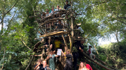 About a dozen children play inside, on top of and around a large treehouse built out of logs and wood planks. It is inside a canopy of trees.
