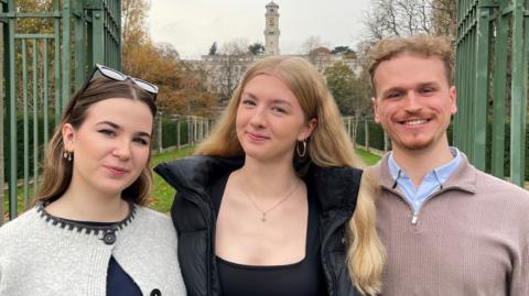 Students Lily-Rose Marsden, Charlie Blair and Max Hegarty standing in front of the gates outside Highfields Park in Nottingham with a university building visible in the distance