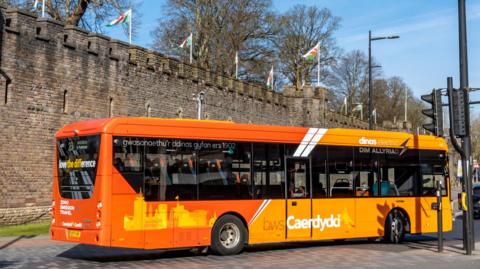 An orange single decker bus in traffic driving past Cardiff Castle.