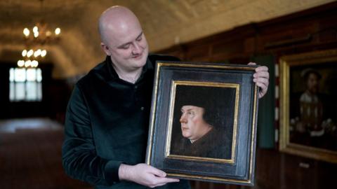 A bald white man wearing a black shirt. He is holding a framed portrait of Thomas Cromwell. The man is stood in a long room with brown wooden walls and a cream ceiling. He is looking down and to the right at the picture he is holding