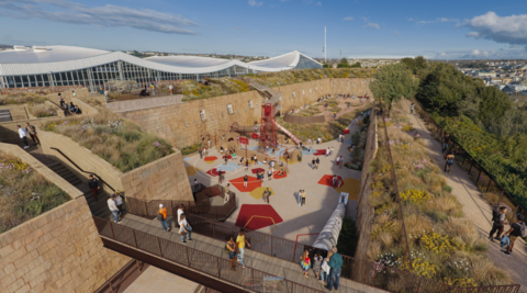 Sunlit outdoor play and leisure area set within a large, terraced stone enclosure. People walk along elevated pathways and gather around playground equipment, including climbing structures and slides. The surrounding landscape features greenery, wildflowers, and views of modern glass-roofed buildings and the city beyond.