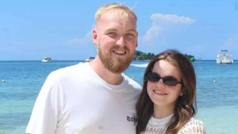 A man with short blond hair and a beard wearing a white T-shirt is next to a shorter woman with long brown hair and a white top. They are standing in front of a blue sea with boats, with a small island in the distance.