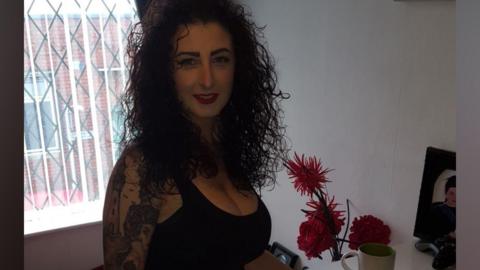 A woman in her 40s, with curly blackhair and wearing a black vest top, smiles at the camera by an upstairs window. She has a sleeve of black and white tattoos on her right arm including an image of a lighthouse and a woman.